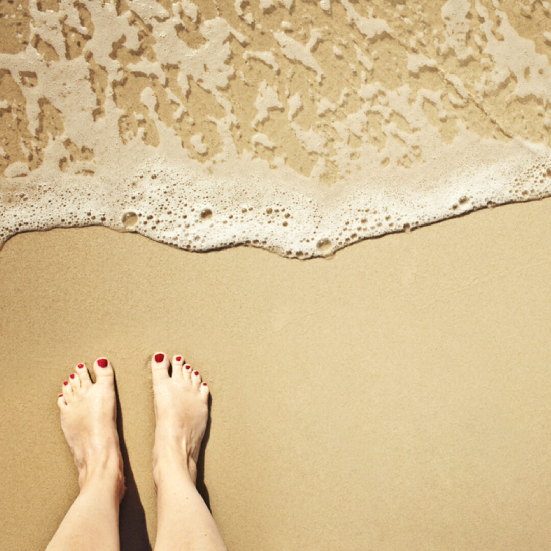 Bare feet with red nail polish on wet sand by gentle ocean waves.