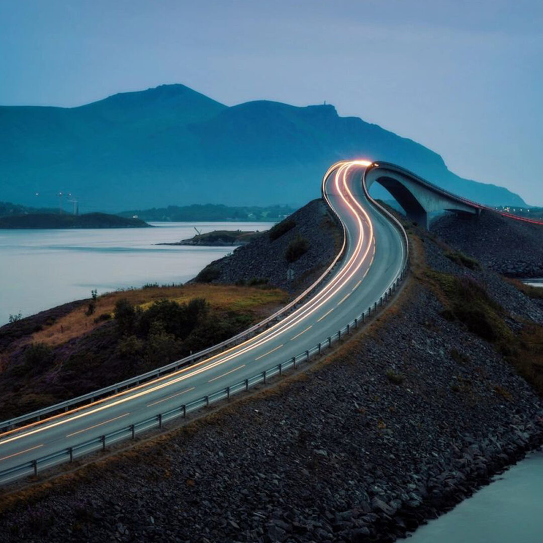 A winding road with light trails against a mountainous backdrop at dusk.