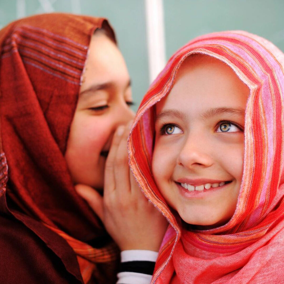 Two girls in headscarves sharing a secret and smiling.